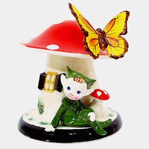 May include: A ceramic figurine of a gnome sitting under a red and white mushroom with a yellow butterfly on top. The gnome is wearing a green outfit and has a white face with blue eyes.