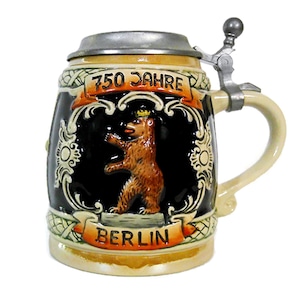 May include: A ceramic beer stein with a hinged metal lid. The stein features a brown bear emblem, the words "750 Jahre" and "Berlin" on banners, and decorative scrollwork. The handle and base are cream-colored.