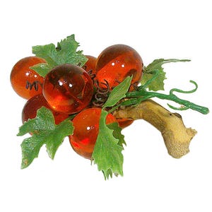 May include: A cluster of orange-red glass grapes with green leaves and a brown stem. The grapes have a glossy finish and are arranged to resemble a bunch. The leaves are a vibrant green, and the stem is a natural brown color.