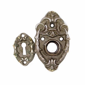 May include: Ornate antique door hardware set, featuring a keyhole cover and a decorative escutcheon. The metal pieces have an aged, tarnished appearance with intricate scrollwork and a central keyhole. The hardware is likely made of cast metal.