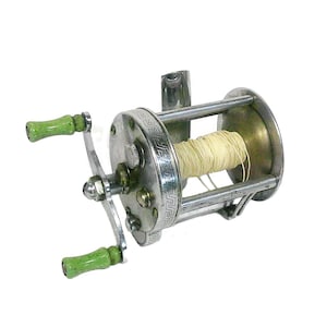 May include: A vintage metal fishing reel featuring a silver-toned cylindrical body with a decorative Greek key pattern along the side plate. It includes a dual-handle crank with two light green speckled knobs and a spool wound with cream-colored line.