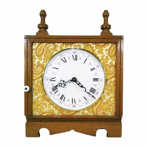 May include: A wooden mantel clock with a square frame and a white clock face featuring black Roman numerals. The clock has a gold paisley pattern background and two decorative finials on top.