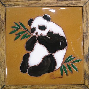 May include: A square ceramic tile featuring a panda bear design. The panda, in black and white, is holding a green leafy branch. The tile is set in a wooden frame with a light brown background.
