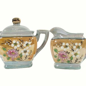 Pode incluir: Um conjunto vintage de a&ccedil;&uacute;car e creme de porcelana com um design floral. O conjunto &eacute; decorado com flores rosa e brancas sobre um fundo azul claro e dourado.