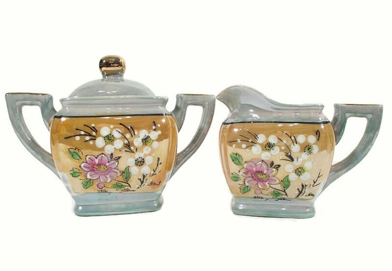 Pode incluir: Um conjunto vintage de a&ccedil;&uacute;car e creme de porcelana com um design floral. O conjunto &eacute; decorado com flores rosa e brancas sobre um fundo azul claro e dourado.