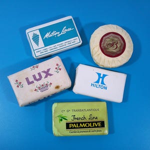 May include: An assortment of vintage soaps on a blue background. The soaps include brands like Matson Lines, Lux, Hilton, and Palmolive. The soaps are in various shapes and sizes, with different packaging designs.