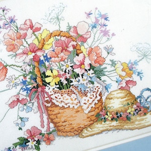 May include: A cross-stitch embroidery of a basket filled with colorful flowers, including pink, yellow, and blue, with a straw hat resting on the side.