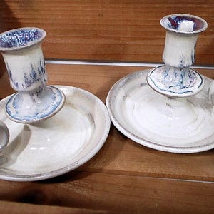 May include: Two white ceramic candle holders with blue and purple accents. Each holder has a small, round base and a larger, round plate with a handle.