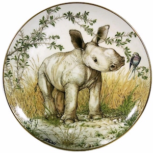 May include: A white ceramic plate with a gold rim features a painting of a baby rhinoceros standing in tall grass. A small blue bird is perched on a branch in the background.