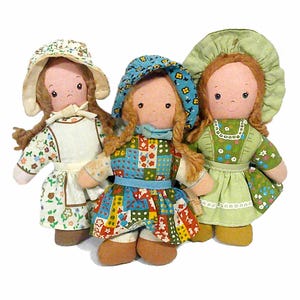 May include: Three fabric dolls with yarn hair and bonnets. The left doll wears a white floral dress and apron. The center doll wears a blue patchwork dress with a matching bonnet. The right doll wears a green floral dress and a green bonnet.