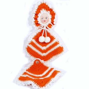 May include: A vintage, handmade crochet doll-shaped kitchen set in orange and white. The set includes a doll-shaped hanging pot holder with a face, two pom-poms, and a matching square pot holder. The set is made of yarn.