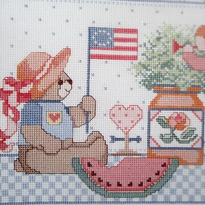 May include: A cross-stitch pattern featuring a teddy bear wearing a red hat and blue shirt, holding a watermelon slice. The bear is sitting next to a jar with a flower and a heart with an arrow. An American flag is in the background.