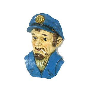 May include: A nautical-themed figurine of a weathered sailor with a blue cap and uniform. The sailor has a beard and a cigarette in his mouth. The figurine is painted in shades of blue, tan, and brown.