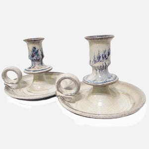 May include: A pair of ceramic candle holders with a white glaze and blue and purple accents. Each holder has a ring handle and a small, round base.