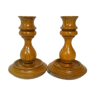 May include: Two wooden candle holders with a warm, golden-brown finish. Each holder has a round base, a central stem with a bulbous shape, and a top designed to hold a candle. The wood grain is visible.