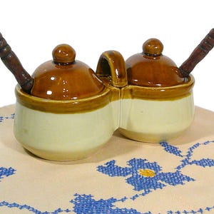 May include: A two-section ceramic condiment server with brown lids and wooden spoons. The server has a cream-colored base with a brown rim and handle. It sits on a cream-colored cloth with blue floral embroidery.