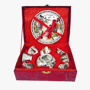 May include: A red velvet box with a white porcelain tea set. The set includes a teapot, six cups, and a plate with a floral design. The box is closed and tied with a red ribbon.