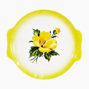 May include: A round, white serving plate with a yellow rim and handles. The plate features a hand-painted yellow hibiscus flower with green leaves. The plate is a decorative piece for serving food.