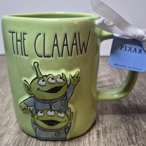 May include: A light green ceramic mug featuring the embossed text THE CLAAAW and a raised graphic of two three-eyed green aliens in blue space suits. A white ribbon is tied to the handle with a blue tag that reads PIXAR and Disney Pixar.