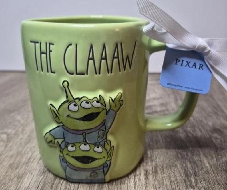 May include: A light green ceramic mug featuring the embossed text THE CLAAAW and a raised graphic of two three-eyed green aliens in blue space suits. A white ribbon is tied to the handle with a blue tag that reads PIXAR and Disney Pixar.