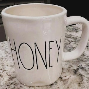 Puede incluir: Una taza de cerámica blanca con un asa curvada. La palabra "HONEY" está impresa en negro, con letras en negrita en la parte delantera de la taza. La taza tiene un acabado mate ligeramente desgastado.
