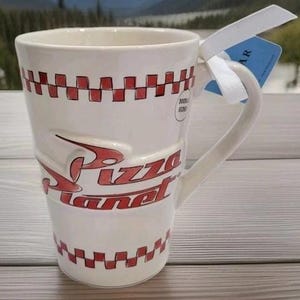 May include: A tall, off-white ceramic mug featuring the red Pizza Planet logo in raised lettering. The mug is decorated with a red and white checkered pattern around the top and bottom rims and includes a white ribbon tied to the handle with a blue tag.