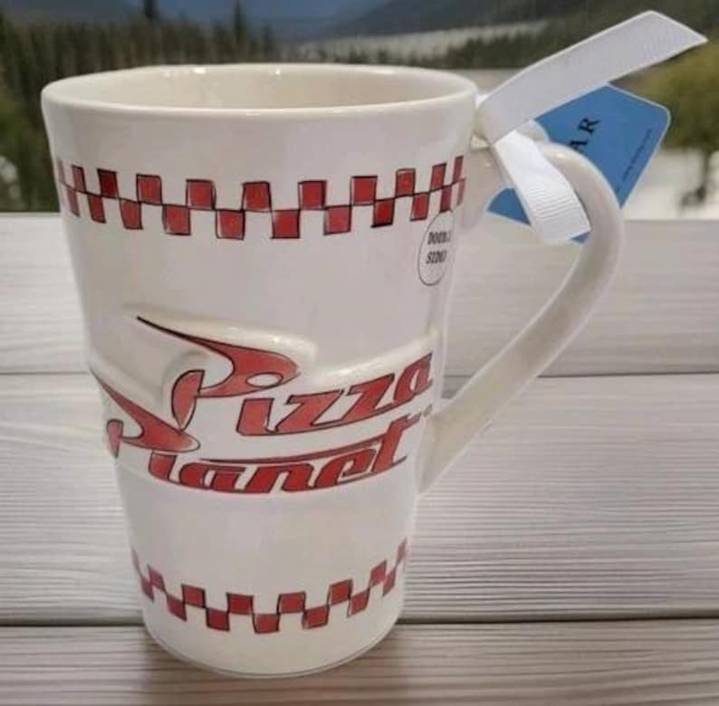 May include: A tall, off-white ceramic mug featuring the red Pizza Planet logo in raised lettering. The mug is decorated with a red and white checkered pattern around the top and bottom rims and includes a white ribbon tied to the handle with a blue tag.