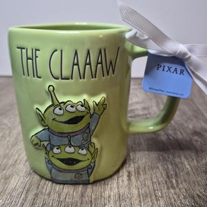 Peut inclure: Un mug en c&eacute;ramique vert clair orn&eacute; de deux extraterrestres verts &agrave; trois yeux en combinaison spatiale, avec le texte THE CLAAAW en relief. Un ruban blanc est nou&eacute; &agrave; l'anse avec une &eacute;tiquette bleue portant l'inscription PIXAR et le copyright.