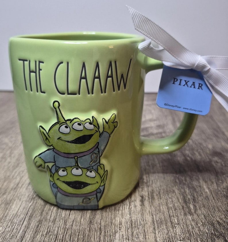 Peut inclure: Un mug en c&eacute;ramique vert clair orn&eacute; de deux extraterrestres verts &agrave; trois yeux en combinaison spatiale, avec le texte THE CLAAAW en relief. Un ruban blanc est nou&eacute; &agrave; l'anse avec une &eacute;tiquette bleue portant l'inscription PIXAR et le copyright.