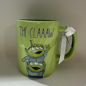 May include: A lime green ceramic mug featuring a raised graphic of two three-eyed aliens in blue space suits. The text THE CLAAAW is printed in black above the aliens. A white ribbon is tied around the handle of the mug, which has a glossy finish.
