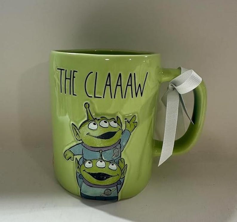 May include: A lime green ceramic mug featuring a raised graphic of two three-eyed aliens in blue space suits. The text THE CLAAAW is printed in black above the aliens. A white ribbon is tied around the handle of the mug, which has a glossy finish.