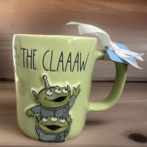 Peut inclure: Un mug en c&eacute;ramique vert clair avec le texte noir THE CLAAAW et un motif en relief repr&eacute;sentant deux extraterrestres verts &agrave; trois yeux en combinaison spatiale grise. Un ruban blanc est nou&eacute; &agrave; l'anse avec une petite &eacute;tiquette bleue.