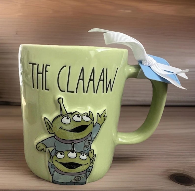 Peut inclure: Un mug en c&eacute;ramique vert clair avec le texte noir THE CLAAAW et un motif en relief repr&eacute;sentant deux extraterrestres verts &agrave; trois yeux en combinaison spatiale grise. Un ruban blanc est nou&eacute; &agrave; l'anse avec une petite &eacute;tiquette bleue.