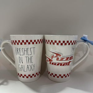 May include: Two white ceramic mugs featuring a red and white checkered border at the top and bottom. One mug displays the black text "FRESHEST IN THE GALAXY" in a handwritten font, while the other features the red "Pizza Planet" logo in relief.