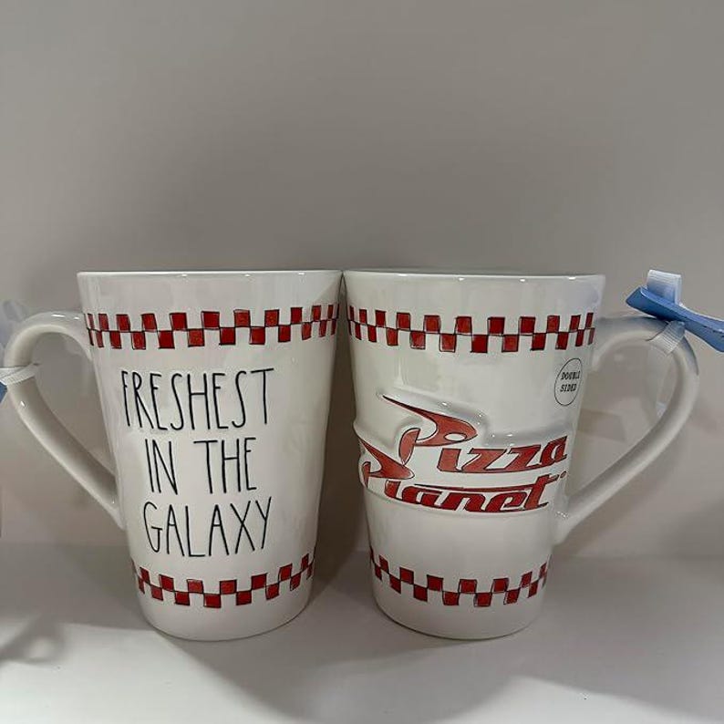 May include: Two white ceramic mugs featuring a red and white checkered border at the top and bottom. One mug displays the black text "FRESHEST IN THE GALAXY" in a handwritten font, while the other features the red "Pizza Planet" logo in relief.