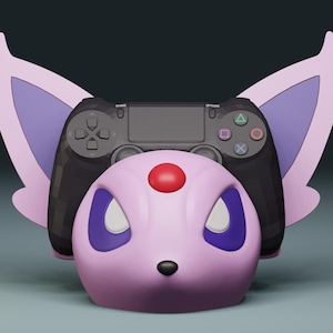 Könnte beinhalten: Ein rosa-lila Controller-Ständer im Espeon-Design mit einem schwarzen PlayStation-Controller, der darauf ruht. Der Ständer hat große Ohren und einen roten Edelstein. Der Controller hat farbige Tasten.