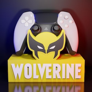 Könnte beinhalten: Ein PlayStation 5-Controller liegt auf einem gelben Block mit dem Wort "WOLVERINE" in Weiß. Der Block zeigt ein Wolverine-Maskenlogo. Der Controller hat weiße Seiten und eine schwarze Mitte.