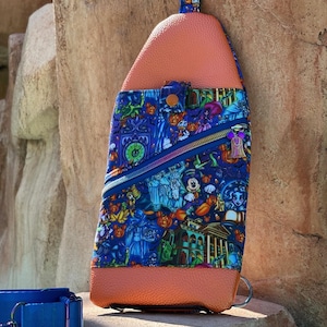 Może przedstawiać: Niebiesko-pomarańczowa torba na ramię z kolorowym nadrukiem przedstawiającym postacie z kresk&oacute;wek i projekt nawiedzonego domu. Torba posiada kieszeń zapinaną na zamek i g&oacute;rny uchwyt. Niebieska obroża dla psa znajduje się na pierwszym planie.