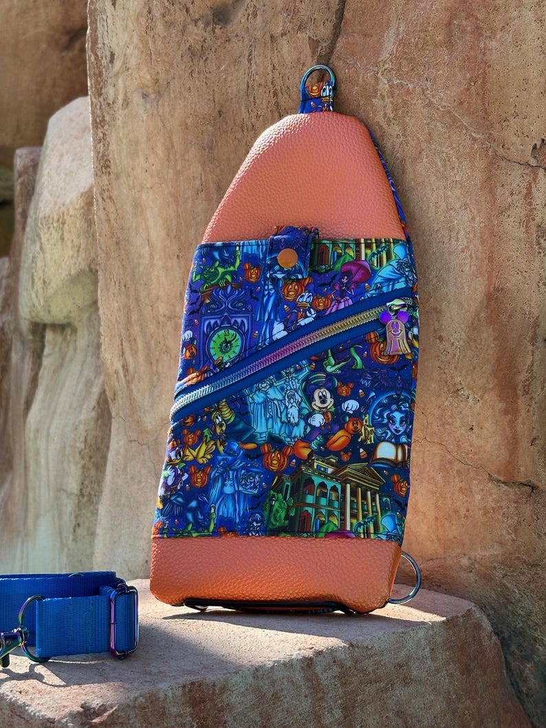 Może przedstawiać: Niebiesko-pomarańczowa torba na ramię z kolorowym nadrukiem przedstawiającym postacie z kresk&oacute;wek i projekt nawiedzonego domu. Torba posiada kieszeń zapinaną na zamek i g&oacute;rny uchwyt. Niebieska obroża dla psa znajduje się na pierwszym planie.