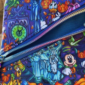 Może przedstawiać: Niebieski przedmiot z tkaniny z nadrukiem o tematyce Halloween z postaciami z kresk&oacute;wek, dyniami i nietoperzami. Projekt obejmuje zegar, duchy i zamek błyskawiczny. Przedmiot ma pomarańczowy guzik i niebieski zamek błyskawiczny.