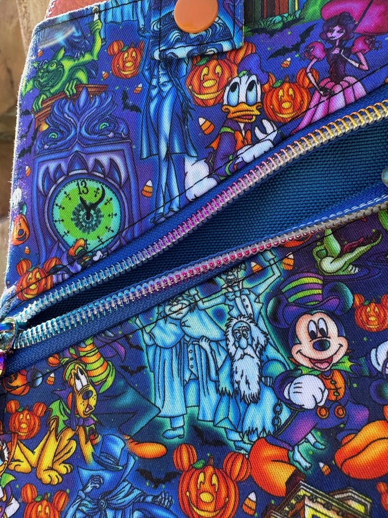 Może przedstawiać: Niebieski przedmiot z tkaniny z nadrukiem o tematyce Halloween z postaciami z kresk&oacute;wek, dyniami i nietoperzami. Projekt obejmuje zegar, duchy i zamek błyskawiczny. Przedmiot ma pomarańczowy guzik i niebieski zamek błyskawiczny.