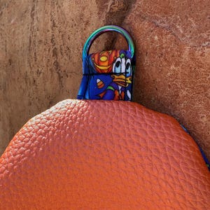 Może przedstawiać: Pomarańczowa, teksturowana torebka z kolorowym paskiem z postacią z kresk&oacute;wki. Pasek ma tęczowy metalowy pierścień. Torebka wykonana jest z materiału przypominającego sk&oacute;rę.