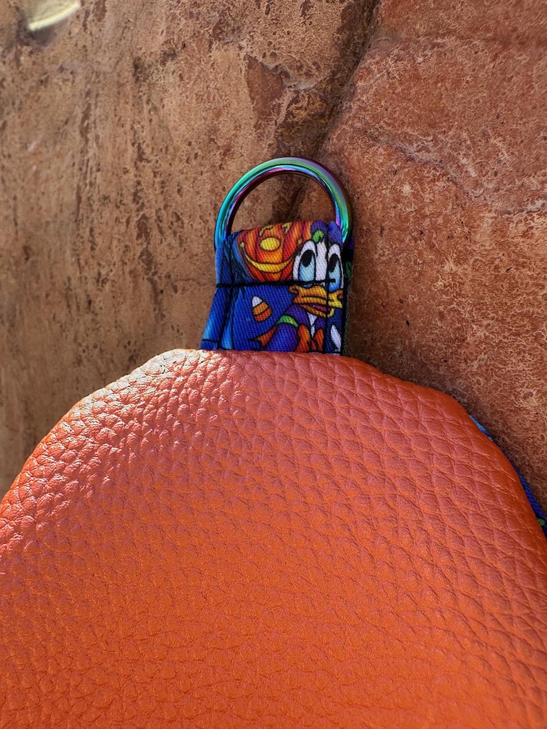 Może przedstawiać: Pomarańczowa, teksturowana torebka z kolorowym paskiem z postacią z kresk&oacute;wki. Pasek ma tęczowy metalowy pierścień. Torebka wykonana jest z materiału przypominającego sk&oacute;rę.