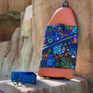 Może przedstawiać: Niebiesko-pomarańczowa torba na ramię z kolorowym nadrukiem przedstawiającym postacie z kresk&oacute;wek i motywem nawiedzonego domu. Torba posiada kieszeń zapinaną na zamek i niebieski pasek z metalowym zapięciem.