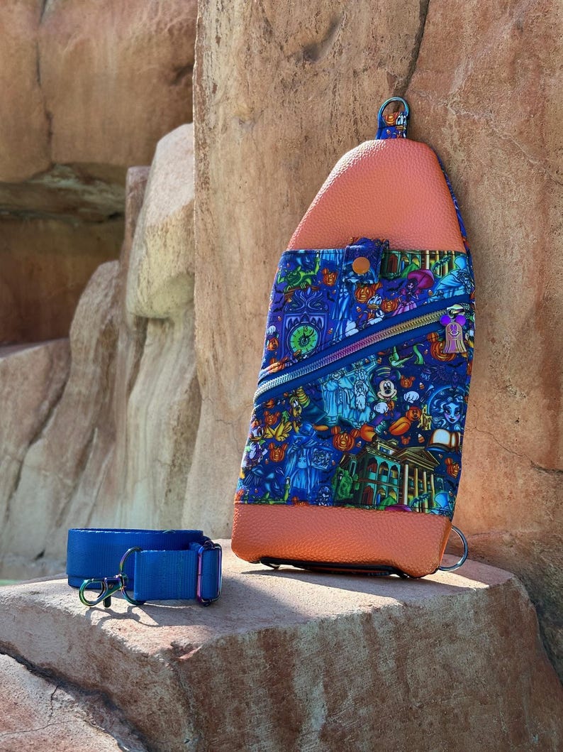 Może przedstawiać: Niebiesko-pomarańczowa torba na ramię z kolorowym nadrukiem przedstawiającym postacie z kresk&oacute;wek i motywem nawiedzonego domu. Torba posiada kieszeń zapinaną na zamek i niebieski pasek z metalowym zapięciem.