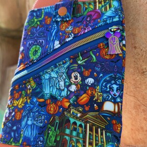 Może przedstawiać: Niebieska torba na ramię z kolorowym nadrukiem o tematyce Halloween, przedstawiającym Myszka Miki, dynie i obrazy nawiedzonych dom&oacute;w. Torba ma niebieski zamek błyskawiczny, fioletowy uchwyt zamka błyskawicznego w kształcie ducha oraz pomarańczowy sk&oacute;rzany pasek i tył.