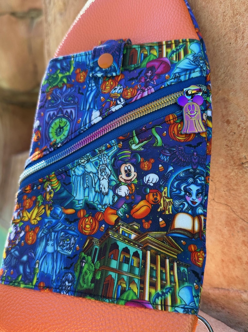 Może przedstawiać: Niebieska torba na ramię z kolorowym nadrukiem o tematyce Halloween, przedstawiającym Myszka Miki, dynie i obrazy nawiedzonych dom&oacute;w. Torba ma niebieski zamek błyskawiczny, fioletowy uchwyt zamka błyskawicznego w kształcie ducha oraz pomarańczowy sk&oacute;rzany pasek i tył.