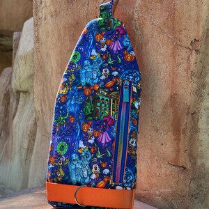 Może przedstawiać: Niebieska, zapinana na zamek torba z kolorowym wzorem o tematyce Halloween, przedstawiającym postacie z kresk&oacute;wek, dynie i nawiedzony dom. Posiada pomarańczowy pasek i niebieski zamek błyskawiczny z r&oacute;żowym suwakiem.