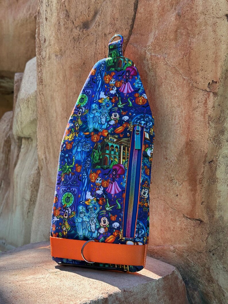 Może przedstawiać: Niebieska, zapinana na zamek torba z kolorowym wzorem o tematyce Halloween, przedstawiającym postacie z kresk&oacute;wek, dynie i nawiedzony dom. Posiada pomarańczowy pasek i niebieski zamek błyskawiczny z r&oacute;żowym suwakiem.