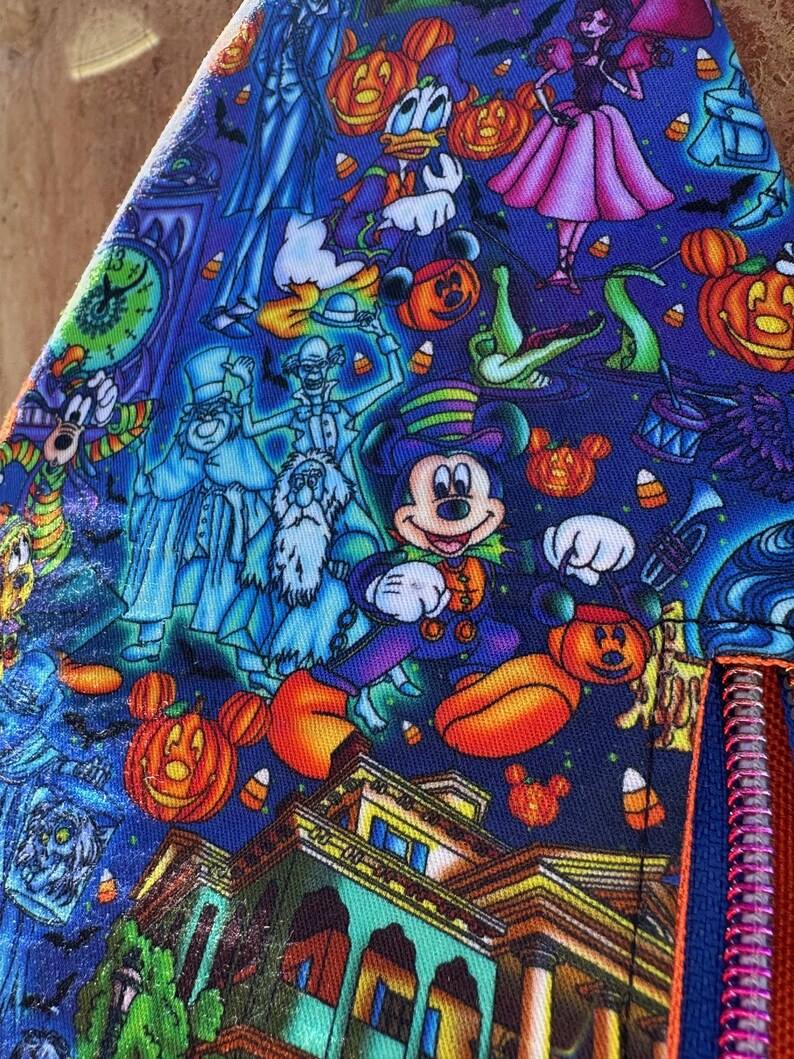 Może przedstawiać: Niebieska tkanina z nadrukiem o tematyce Halloween z Myszka Miki, Kaczorem Donaldem i innymi postaciami. Projekt zawiera dynie, duchy i nawiedzony dom. Widoczny jest czerwony zamek błyskawiczny.